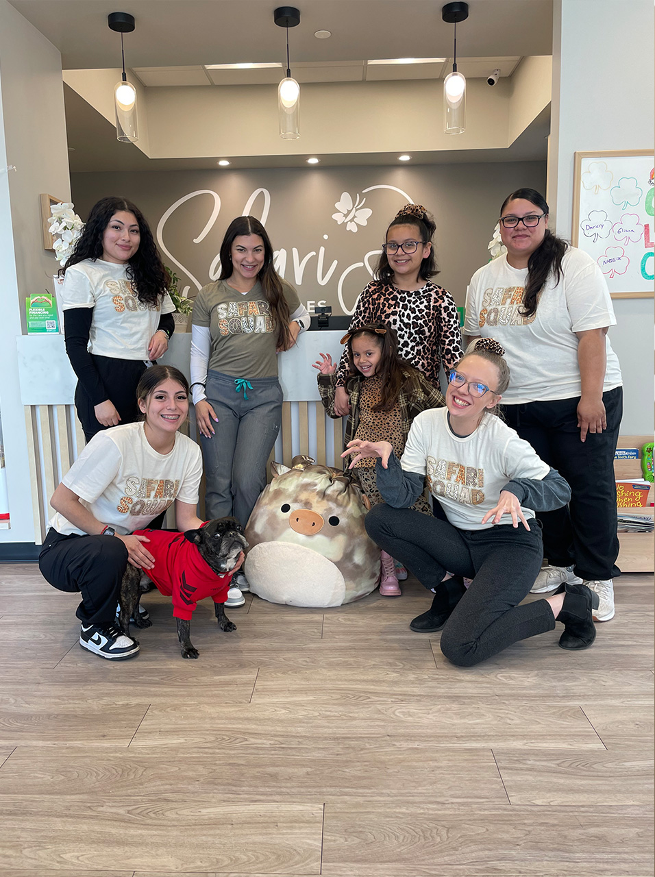 The image shows a group of people posing together in front of a storefront, with one person holding a small dog and another person kneeling on the floor. They are all smiling and appear to be enjoying themselves.