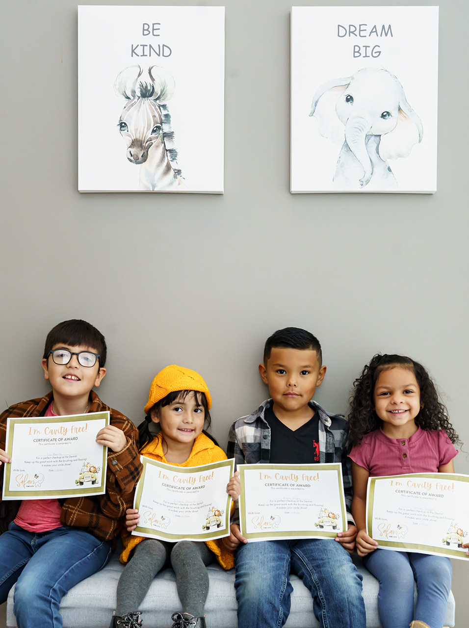 The image shows a group of four children, likely boys, standing in front of a wall with two framed pictures. They are holding certificates and appear to be receiving awards, as indicated by the text on the certificates.