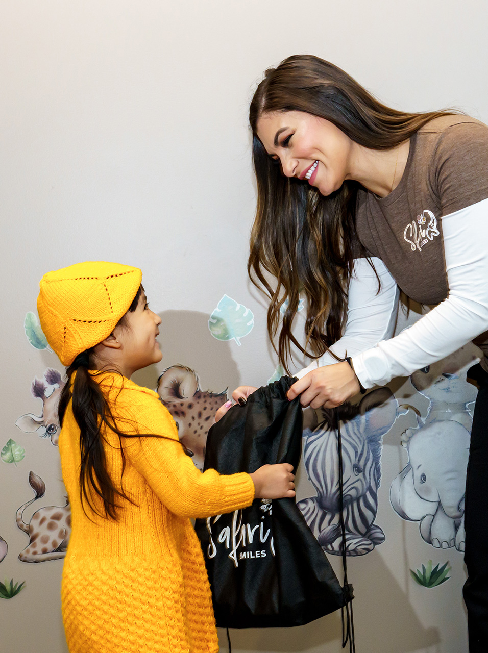 The image shows a woman in a brown shirt and a child wearing a yellow jacket, both standing near a wall with a cartoon animal on it. They are handing over what appears to be a black bag.