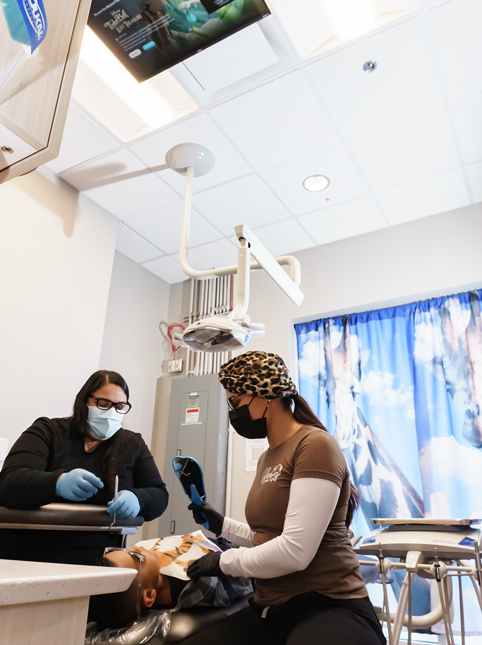 A medical professional wearing a face mask and surgical gown is attending to a patient in a dental chair, with a TV screen displaying a program in the background.