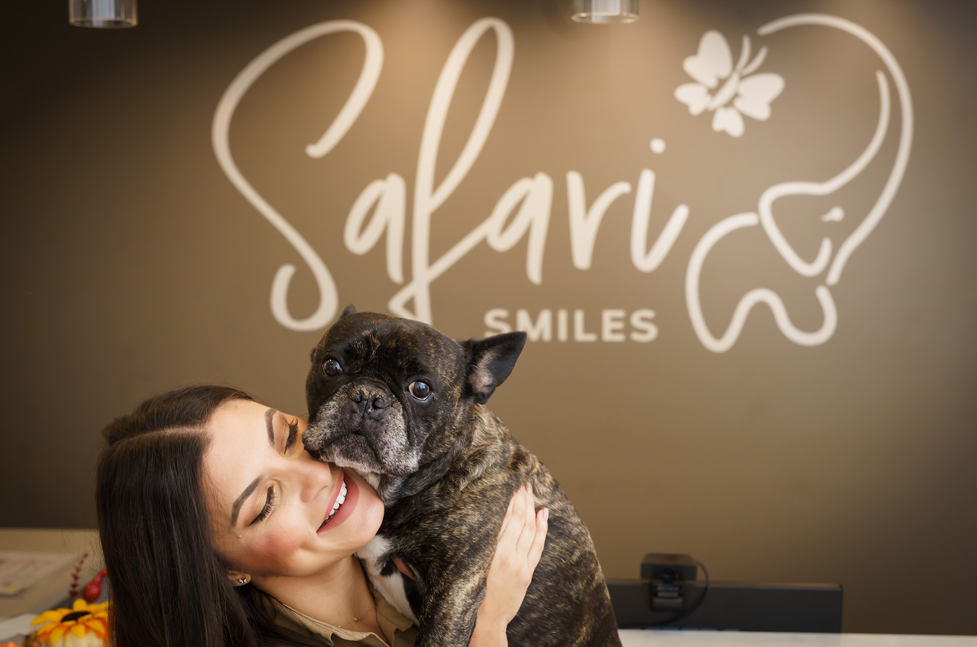 The image shows a woman holding a small dog in front of a sign for  Salvador Smiles.