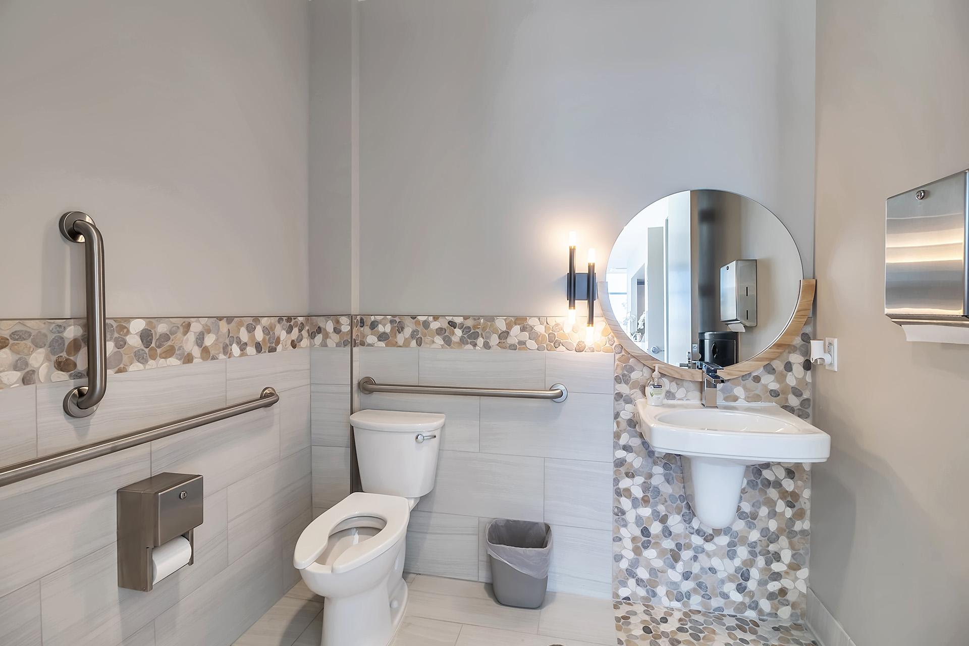 The image shows a well-lit, modern bathroom with white walls and a tiled floor. It features a toilet, sink, mirror, and a glass door shower enclosure with a mosaic backsplash.