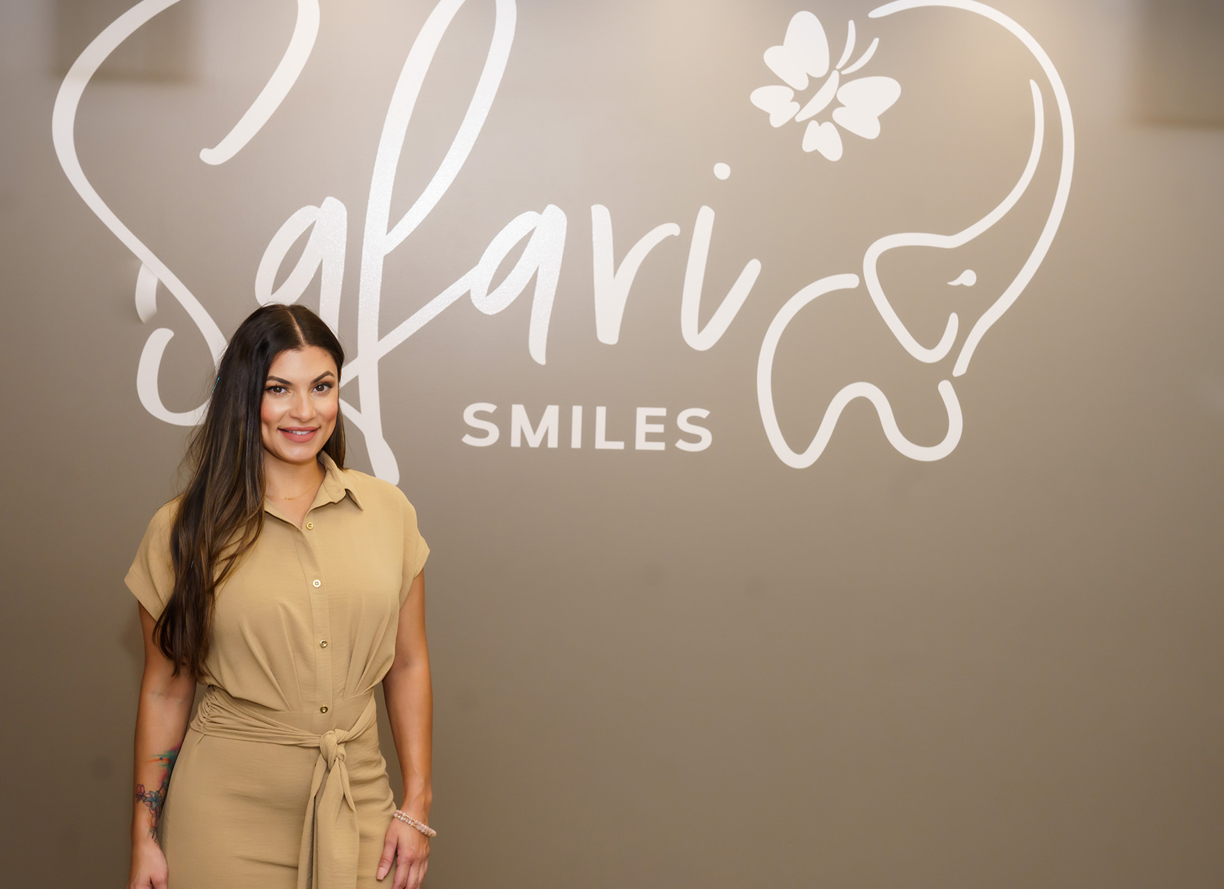 The image shows a woman standing in front of a sign for 'Salafi Smiles,' which is likely a dental practice. She is wearing a beige blazer and has long hair, and there's a logo with a stylized giraffe head on the sign behind her.