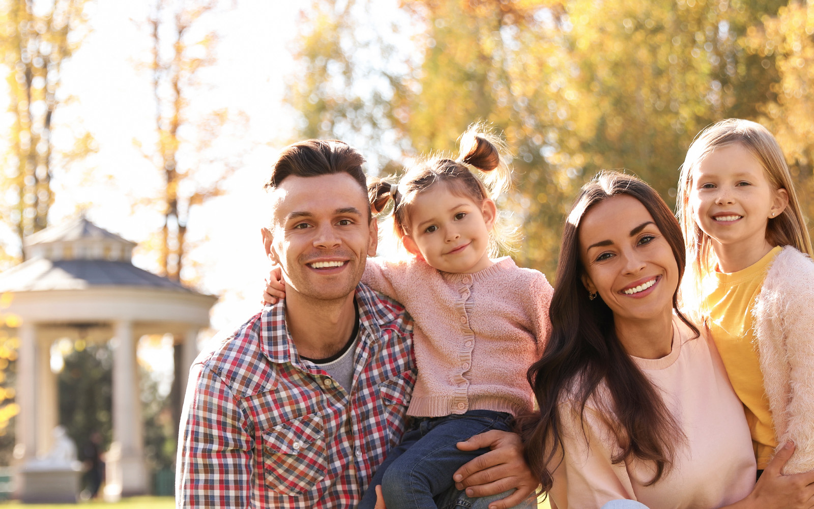 A family of four, including a man and woman with two children, posing for a photo in an autumn park setting.