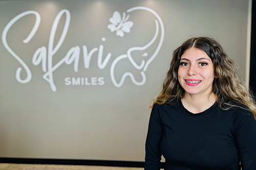 The image shows a woman standing in front of a sign with the word  Salavar  and a logo that appears to be related to dental care or orthodontics. She is smiling at the camera, wearing a black top, and has long hair.