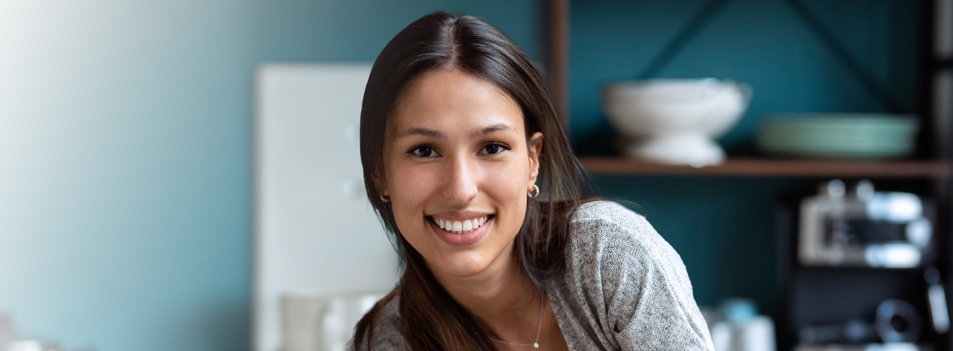 The image shows a woman smiling at the camera, with a blurred background that suggests an indoor setting, possibly a kitchen or dining area.