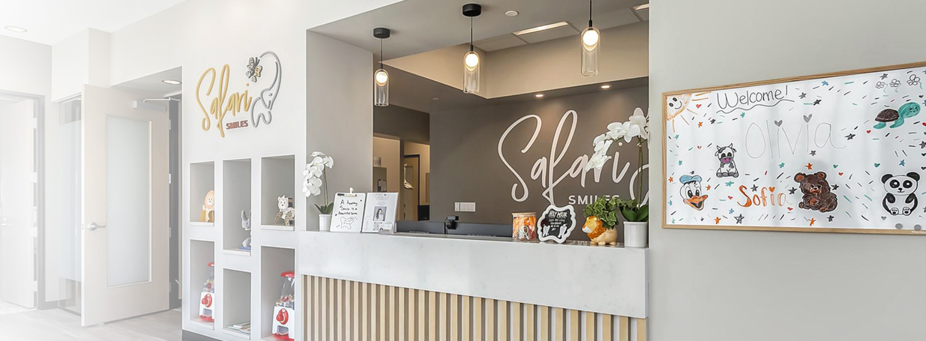 The image shows an interior space, likely a store or boutique, with a modern design featuring a white color scheme and minimalist decor. There is a reception desk in the foreground with a sign that reads  SWEET,  and on the wall behind the desk are various framed items and a large mirror reflecting the room. The floor appears to be made of tiles, and there are decorative elements such as potted plants and framed pictures. The lighting is bright, suggesting an open and airy atmosphere.