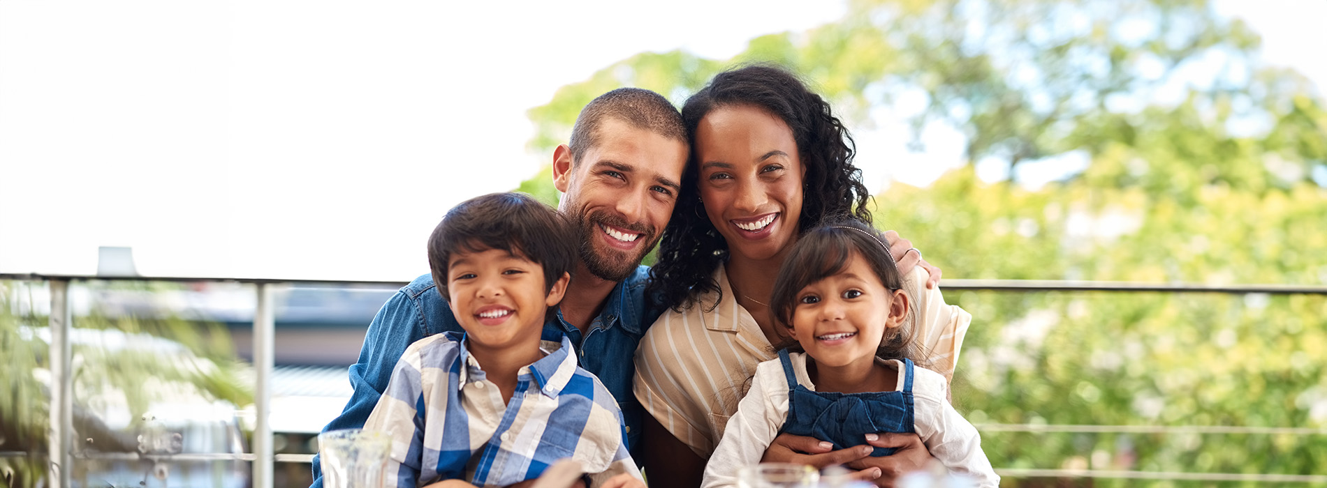 The image shows a family of four, including two adults and two children, posing together outdoors with a building in the background.