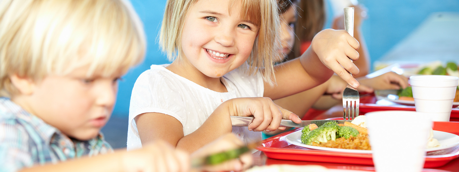 A young girl is smiling and holding a plastic fork with food on it, sitting at a dining table with other children eating.