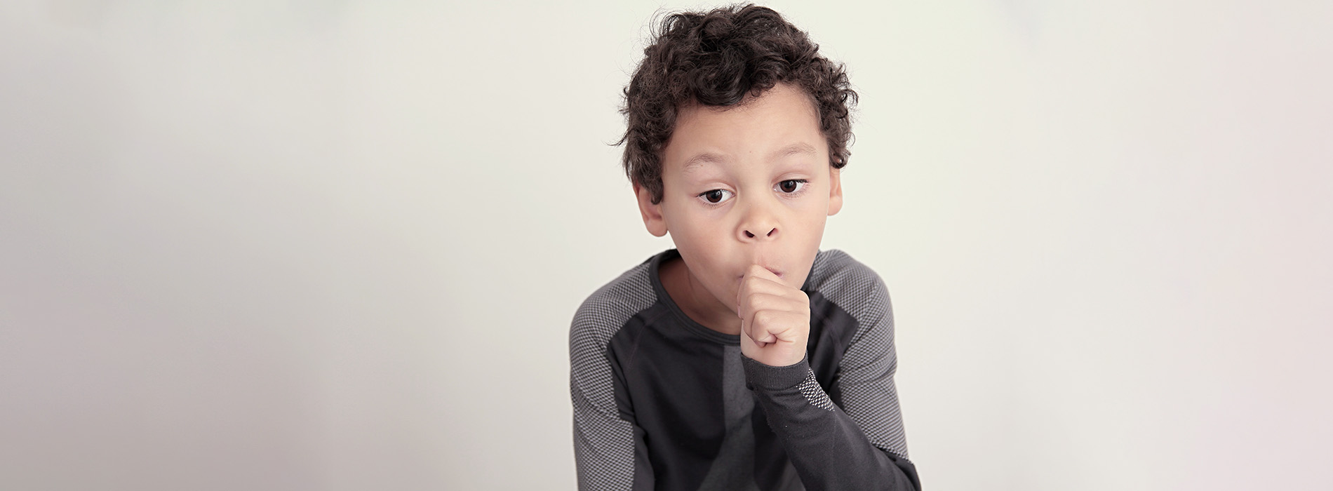 The image shows a young boy with his hand near his mouth, positioned in front of a plain background.