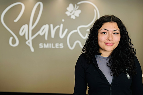 A woman with long brown hair, wearing a black top with white text, stands in front of a sign that reads  Salavador Smiles .