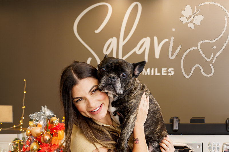 The image shows a woman holding a small dog, both are indoors with a festive atmosphere, and there is a sign in the background that reads 'SALGARI FAMILY DENTISTRY.