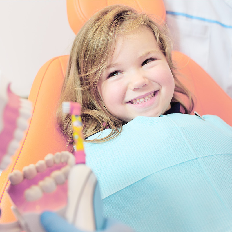 A young girl is sitting in a dental chair, smiling at the camera, with her mouth open for a dental examination.