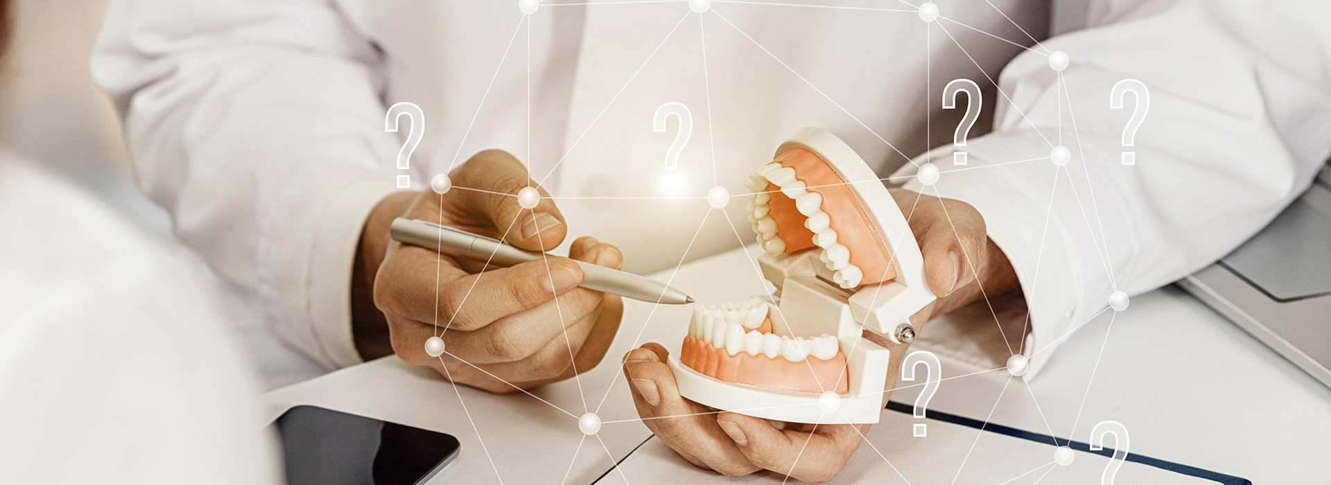 A person s hands holding a model of a human mouth with teeth, set against a background that includes a computer screen displaying medical or dental information.