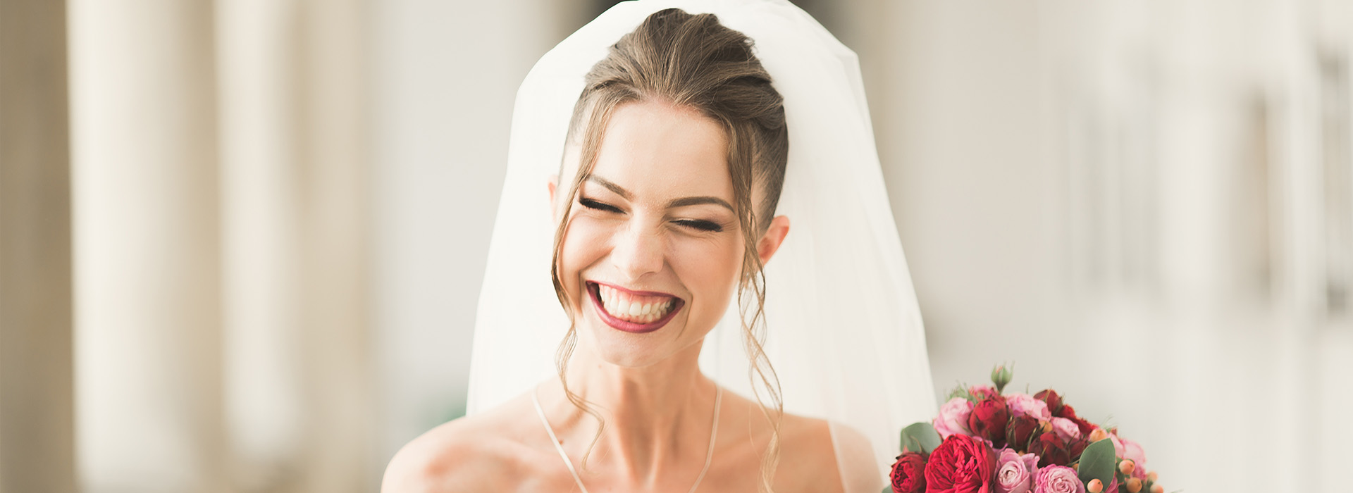 The image is a photograph of a woman in a wedding dress, wearing a necklace and laughing with her mouth open. She is smiling at the camera while holding what appears to be a bouquet of flowers.