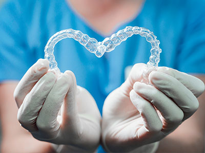 The image shows a person s hands holding a transparent dental retainer shaped like a heart, with gloves on and a blue background.