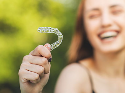 A young woman holding a clear, single-tooth-shaped dental appliance with a smile, against a blurred background of greenery.