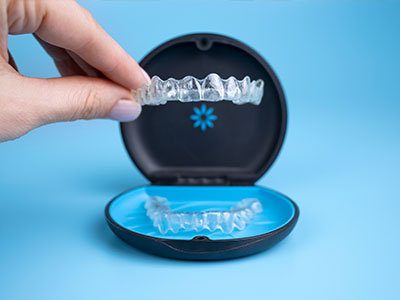 The image shows a person s hand holding an open case containing a clear, transparent dental retainer with a blue base.