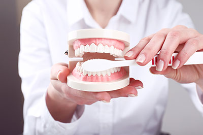 A person s hand holding a dental model with missing teeth, placed in front of a dentist s mouth and instruments.