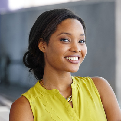 A woman with a radiant smile, wearing a yellow top and standing against a building wall.