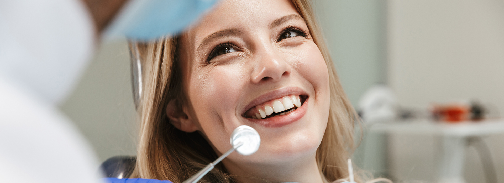 The image shows a woman smiling at the camera while seated in a dental chair, with a dentist visible behind her.