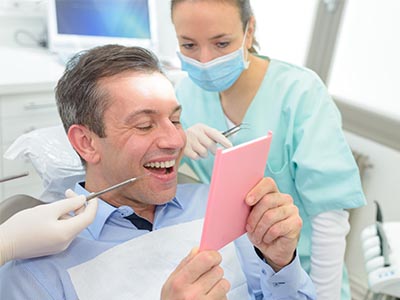 The image shows a man sitting in a dental chair, holding up a pink card with a surprised expression, while a dental professional is standing behind him, smiling and observing the card.