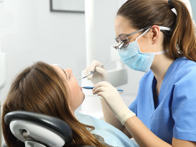 A dental hygienist performing a teeth cleaning procedure on a patient, with the patient seated in an examination chair and the hygienist standing behind, both wearing protective face masks and using dental instruments.