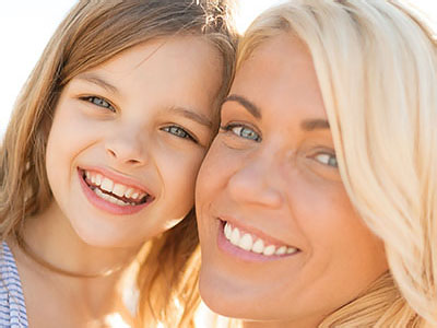 The image shows a woman and a young girl, both smiling, with the woman appearing to be of middle age and the child having light brown hair. They are outdoors during daylight, and the setting suggests a warm climate.