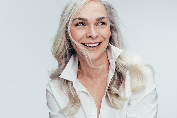 The image shows a woman with short hair, wearing a white top, smiling and looking directly at the camera. She has a neutral background that does not distract from her.