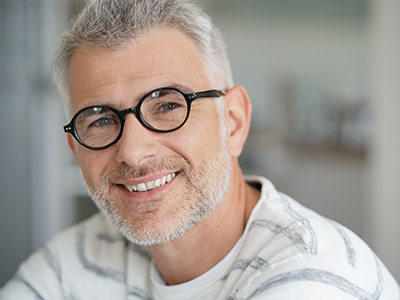 The image features a middle-aged man with glasses, smiling warmly at the camera. He has short grey hair and is wearing a white t-shirt with a patterned overlay.