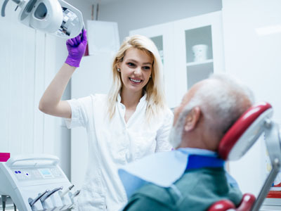 A dental hygienist in a white coat and gloves is assisting an older man with a blue beard, who is seated in a dental chair, receiving dental care.