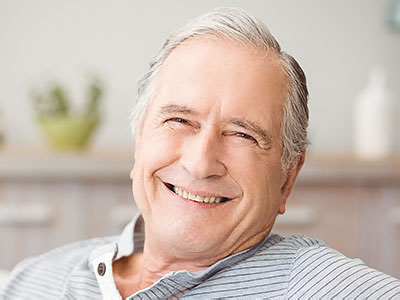 The image is of an older man with a smile, sitting in a chair indoors.