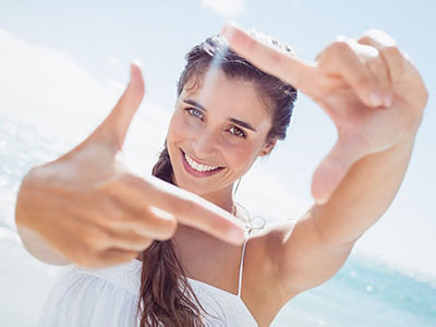 The image shows a smiling woman with long hair, holding up her hand with fingers splayed to create the appearance of a frame around herself. She is outdoors, in front of a clear blue sky and ocean, with sunlight reflecting off the water.