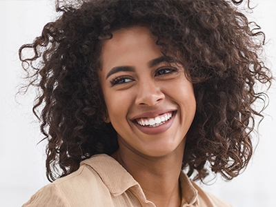 The image shows a smiling woman with curly hair, wearing a white top and a beige blazer. She has her eyes closed and appears to be in a positive mood.